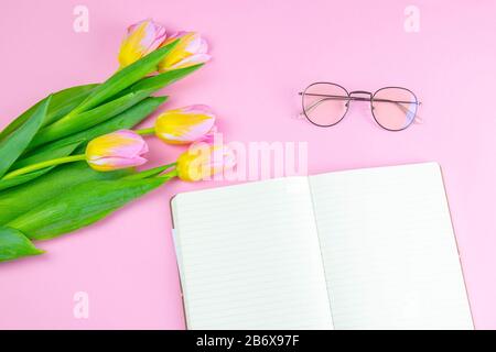 Colorful pinc and yellow tulip with open notebook and eyeglasses flat lay isolated on pinc background top view. Spring flower. Womans day, mothers day Stock Photo