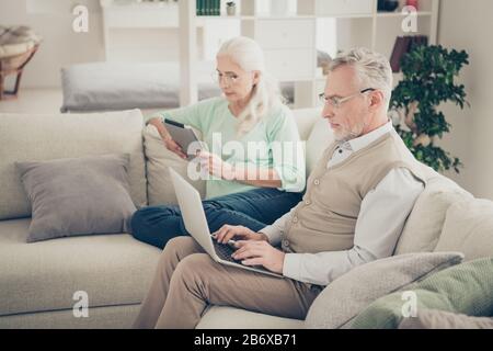 Photo of serious concentrated businessman using netbook sending e-mail and focused calm granny enjoying digital tablet they are sitting on divan Stock Photo