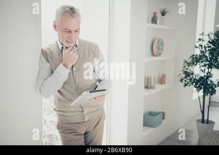 Photo of white haired aged man leaning window indoors reading e-book interesting article Stock Photo