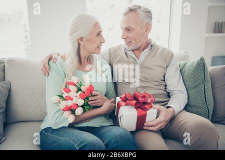 Adorable aged pair sitting comfy sofa celebrating anniversary hold hands red tulips bunch and giftbox Stock Photo