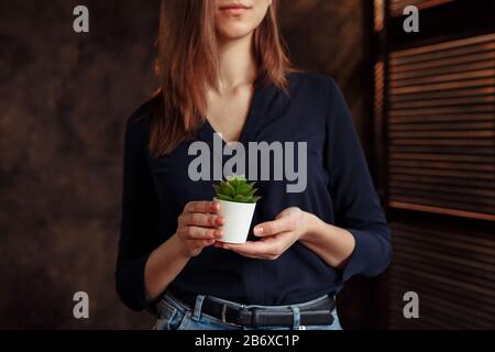 A girl in a blue jacket is holding a white pot with a cactus. Only the girl's smile is visible in the photo. the photo is made in a dark style Stock Photo