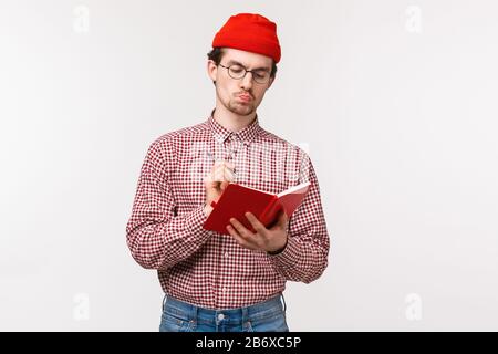 Waist-up portrait of serious-looking bossy male manager in red beanie, glasses, pouting thoughtful while writing down notes in red notebook, making Stock Photo