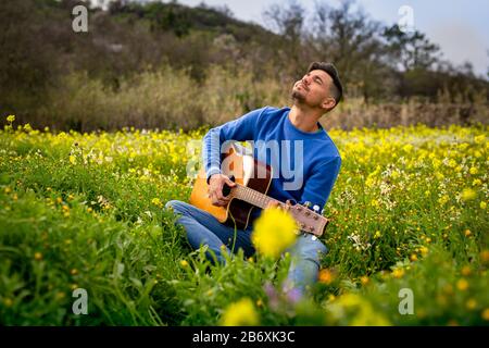 young man sitting in his field of yellow wildflowers, plays the guitar and enjoys Stock Photo