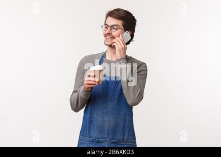 Cheerful friendly young male employee in apron having break, talking on phone and drinking coffee, smiling look away while calling friend discuss Stock Photo
