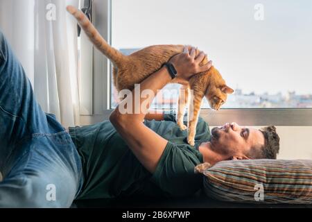 young man lying on a pillow under the window, takes a tabby cat with his hands to kiss him Stock Photo