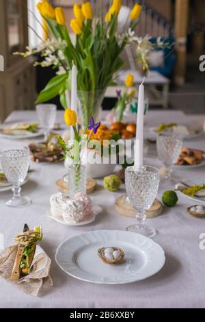 Easter festive spring table setting decoration, eggs in nest, fresh yellow tulips in vase, marshmallows, feathers, family dinner or breakfast concept, Stock Photo
