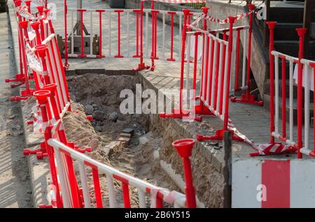 Dangerous pit on the sidewalk surrounded by warning red-and-white barriers. Repair of paving slabs. Stock Photo