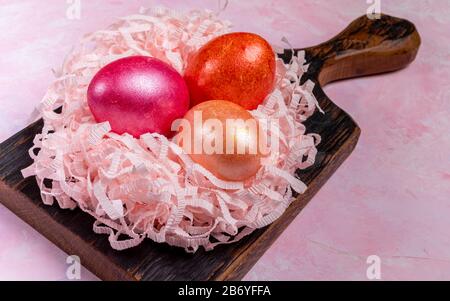 Three bright eggs in nest from pink paper shreds on dark wooden kitchen board on pink textured background. Painted eggs as symbols of Easter. Close-up Stock Photo