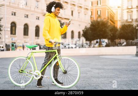 Happy Afro man using mobile smartphone outdoor - Young guy having fun listening music with headphones while riding with bike in city Stock Photo