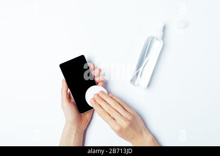 Female's hands wiping mobile phone with alcohol decontaminating agent, top view on white background. Stock Photo