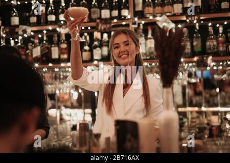 Happy smiling girl raised a toast on the big birthday party. Holding tasty cocktail in a hand. Pretty girl in white jacket, stylish hair lying on the Stock Photo