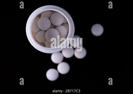 Black background. An open white bottle inside white round tablets. Separate tablets in the blur. View from above. Stock Photo