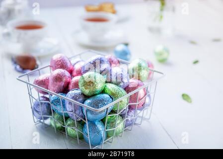 A metal modern basket filled chocolate easter eggs in multicolor foil on a white wooden table with tea cups on background. Minimalist festive spring c Stock Photo