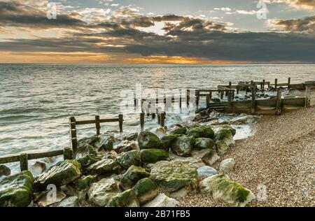 Winter seascape vista of old, battered and broken sea defences with warm glowing sunset on the distant horizon Stock Photo