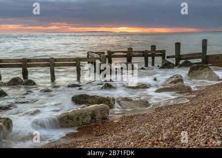 Winter seascape vista of old, battered and broken sea defences with warm glowing sunset on the distant horizon Stock Photo