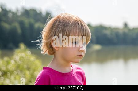 little girl looks thoughtfully out over a lake on a nice summer day. lake and trees in the background. the picture is closeup Stock Photo