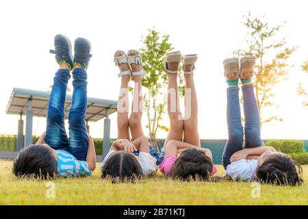 Large group of happy Asian smiling kindergarten kids friends lying on grass and raised leg into sky together in the park on the green grass on sunny s Stock Photo