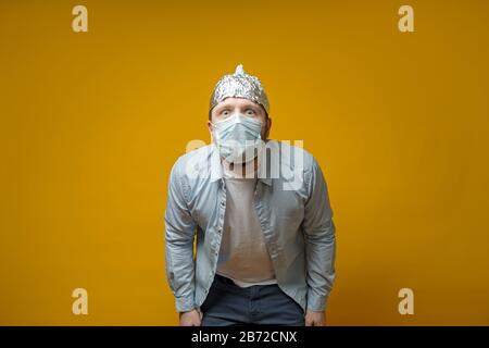 Man in the medical mask is very afraid of contracting the virus, he put on a tin foil hat for protection and looks at the camera.