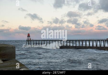 A wave crashes against the structure of the East Pier protecting the entrance to Whitby harbour in North Yorkshire Stock Photo