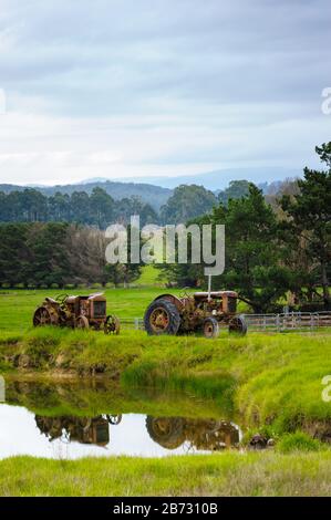 Yarra Valley, Victoria with rolling grassy, wooded farmland and two farm tractors parked on a dam wall with their reflection in the water. Stock Photo