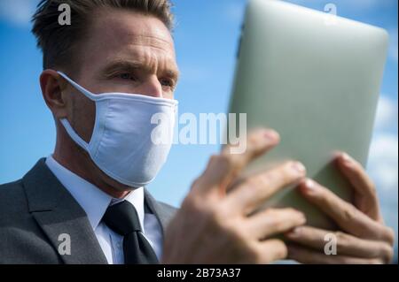 Businessman wearing surgical face mask using his tablet computer outdoors as a mobile office in isolation Stock Photo