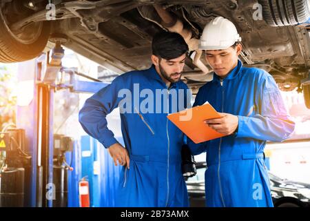 Mechanic in blue work wear uniform inspects the car bottom with his assistant. Automobile repairing service, Professional occupation teamwork.