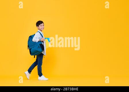 Smiling handsome 10 year-old schoolboy holding books and backpack walking in yellow studio background for education concept Stock Photo