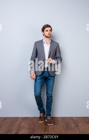 Full-length portrait of young handsome confident man in jacket holding glasses in his hand against gray background. Stock Photo