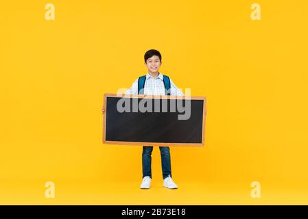 Smiling handsome 10 year-old mixed race boy holding empty blackboard isolated on yellow studio background for education concept Stock Photo