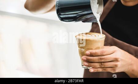 Professional barista pouring steamed milk into coffee glass cup making beautiful latte art Rosetta pattern in cafe Stock Photo