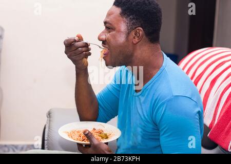 young man sitting in an armchair eating spaghetti holding a plate in his hand. Stock Photo