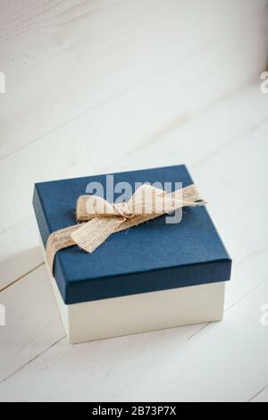 Square gift box with blue lid and burlap bow on white wooden background. Stock Photo