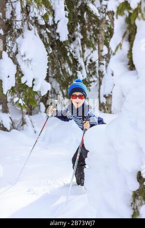 Young boy peeking out from behind heavy snow clad trees, with playful and cheeky expression on his face, tongue out. Dressed in knit wear. Stock Photo