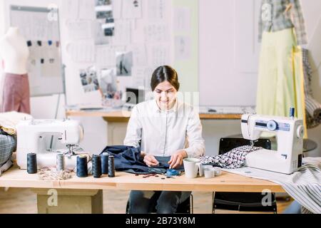 Smiling female fashion designer sitting at office desk. Young woman sewing with sewing machine at studio while sitting at her working place Stock Photo