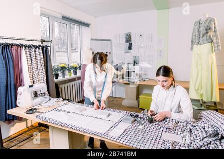 Two pretty women dressmakers working in the fashion atelier with stylish fabric on which they have marked out the pattern with tailors chalk. Checkere Stock Photo