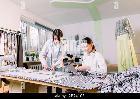 Two pretty women dressmakers working in the fashion atelier with stylish fabric on which they have marked out the pattern with tailors chalk. Checkere Stock Photo