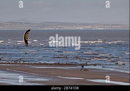 Portobello Beach, Edinburgh, Scotland, UK. 13th Mar 2020. Man Skateboard Kitesurfing along the sandy beach. Sunny but cool, Wind: ENE 18 km/h with wind gusts of 32 km/h, Temperature of 6 degrees centigrade. Stock Photo