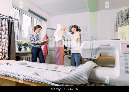 Two female tailors or seamstress taking mannequin measurements with measuring tape in fashion design studio, woman designer working with sketches in w Stock Photo