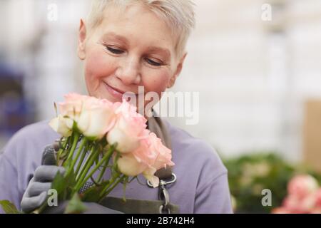 Mature gardener with short hair holding roses in her hands and smelling them Stock Photo
