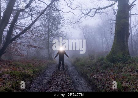 A spooky, hooded figure with a bright light coming from their head. In a forest on a foggy winters day. Stock Photo