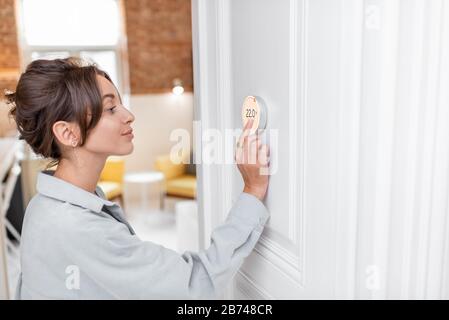 Woman regulating heating temperature with a modern wireless thermostat installed on the white wall at home. Smart home heating regulation concept Stock Photo