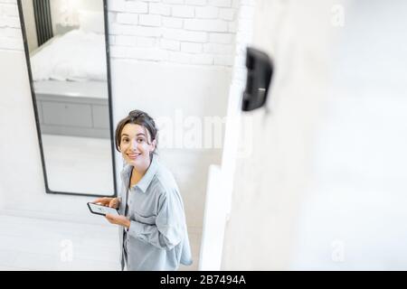 Woman controlling alarm system with a smart phone wireless, standing in the room with motion sensor mounted on the wall Stock Photo
