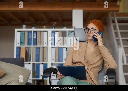 Young serious businesswoman in eyeglasses sitting on chair with document and talking on mobile phone at office Stock Photo
