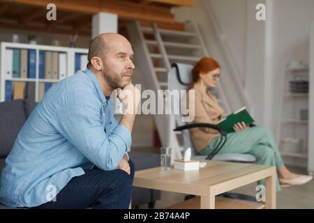 Young pensive man sitting on sofa and thinking about something with young businesswoman sitting in the background at office Stock Photo