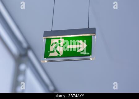 Close up of illuminated emergency exit sign (colored green and white). Modern design, with running figure and arrow to the right. Neutral, background.