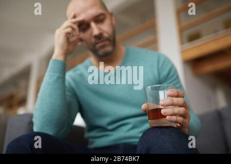 Man sitting on sofa and looking at glass with whiskey in his hand he drinking during his problems Stock Photo