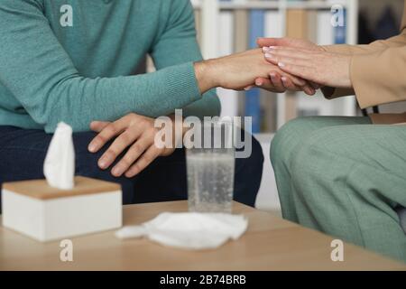 Close-up of woman holding hands of the man and calming him while they sitting on sofa during psychology therapy Stock Photo