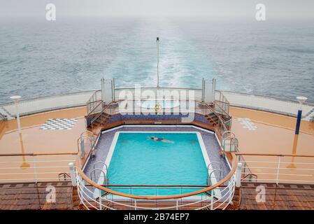 Crossing the Atlantic Ocean from Brooklyn to Southampton onboard the ocean liner Queen Mary 2. A lonely swimmer in the swimming pool on the aft main deck. Stock Photo