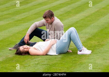 Beautiful pregnant young woman lying down on the grass with her partner sitting next to her Stock Photo