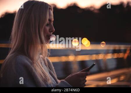 Young woman blowing bubble gum while using smart phone sitting on bench at dusk Stock Photo